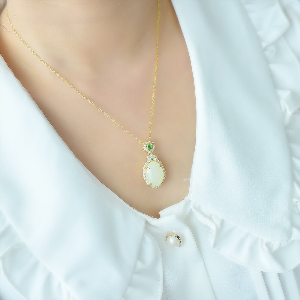 An oval Hetian jade main stone, topped with a golden floral crown, and adorned with emerald green gemstones; a pendant necklace.
